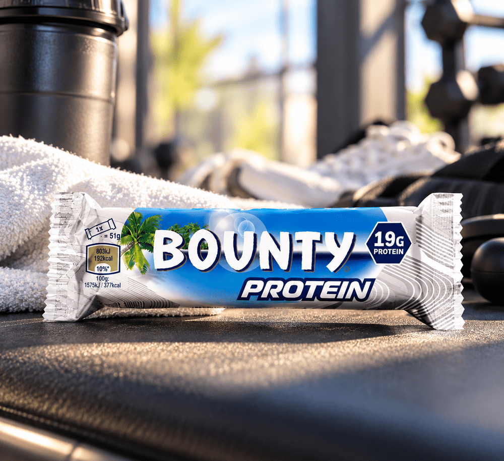 Barre Bounty Protein contenant 19 g de protéines, emballage bleu et blanc posé sur un banc de sport avec accessoires de musculation.