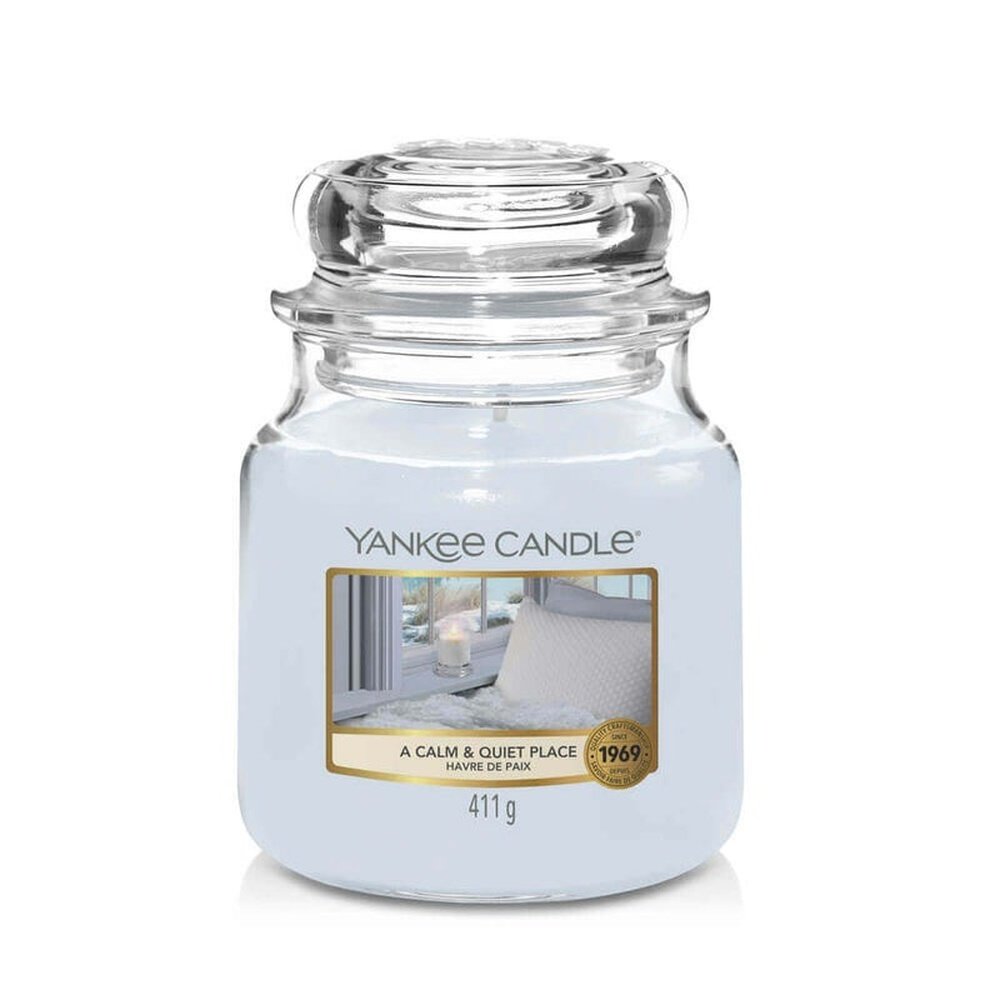 Un emballage sur fond blanc avec une bougie gris clair dans un bocal en verre, un capuchon en verre et une étiquette avec une bougie blanche devant une fenêtre et un lit douillet