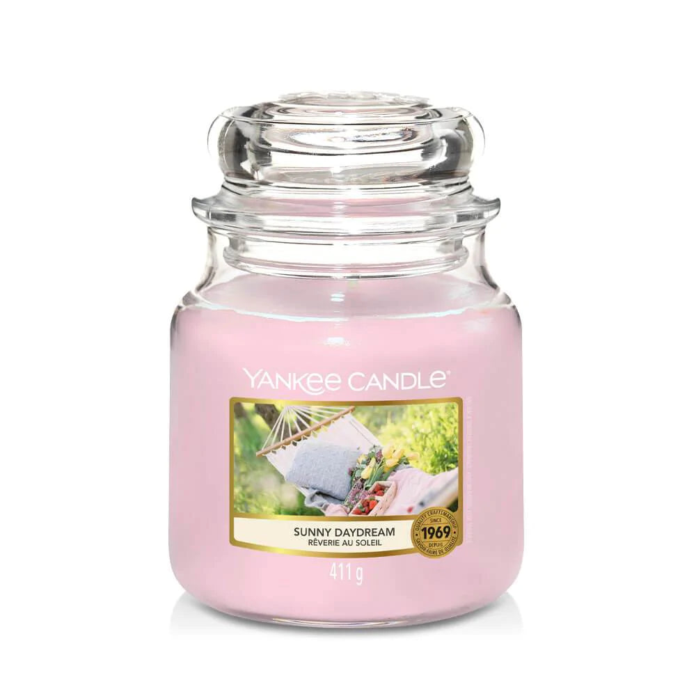 Un emballage sur fond blanc avec une bougie rose dans un bocal en verre, un capuchon en verre et une étiquette avec des tissus et des fleurs sur un hamac blanc dans un jardin