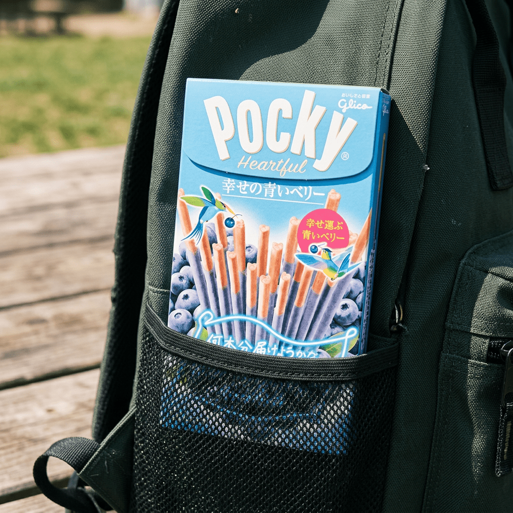 Le paquet de bâtonnets chocolatés, Glico Pocky Heartful Blueberry est dans une poche fillet sur le coté d’un sac à dos noir – disponible sur My American Shop