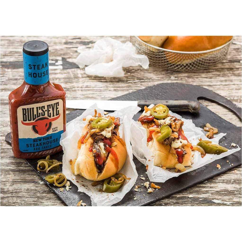 Une bouteille rouge avec les étiquettes bleues et 2 hot dogs avec de la viande, de la sauce et des jalapenos verts sur une planche noir sur une table en bois