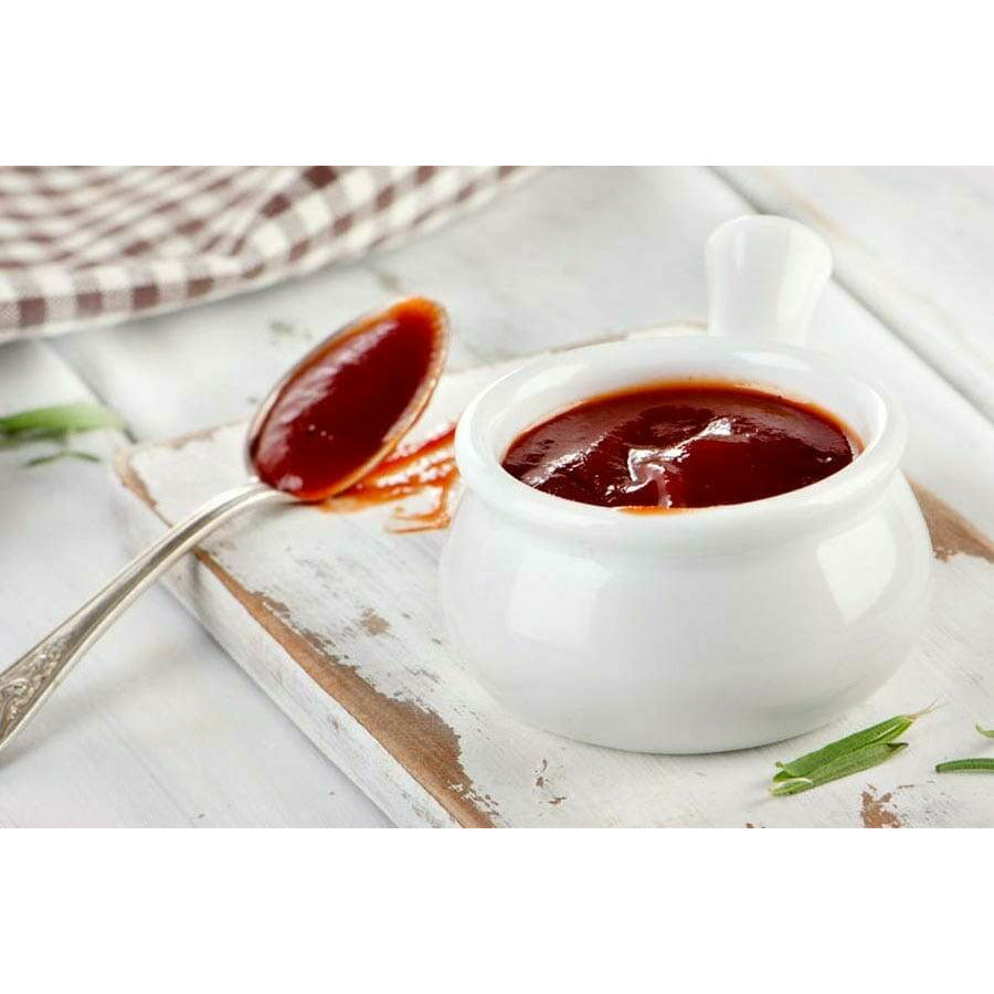 Un récipient en verre blanc rempli d’une sauce rouge et à côté une cuillère avec de la sauce sur une table en bois peinte en blanc 