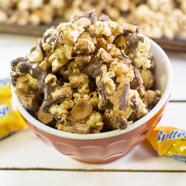 Un bol rouge de popcorn au chocolat avec autour des paquets jaunes de Butterfinger sur une table blanche