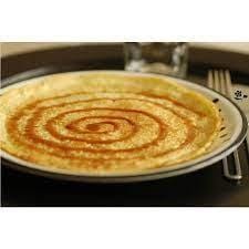 Une crêpe dans une assiette avec du sirop brun en spiral et sur le coté droit une fourchette et un verre, le tout sur un plateau noir