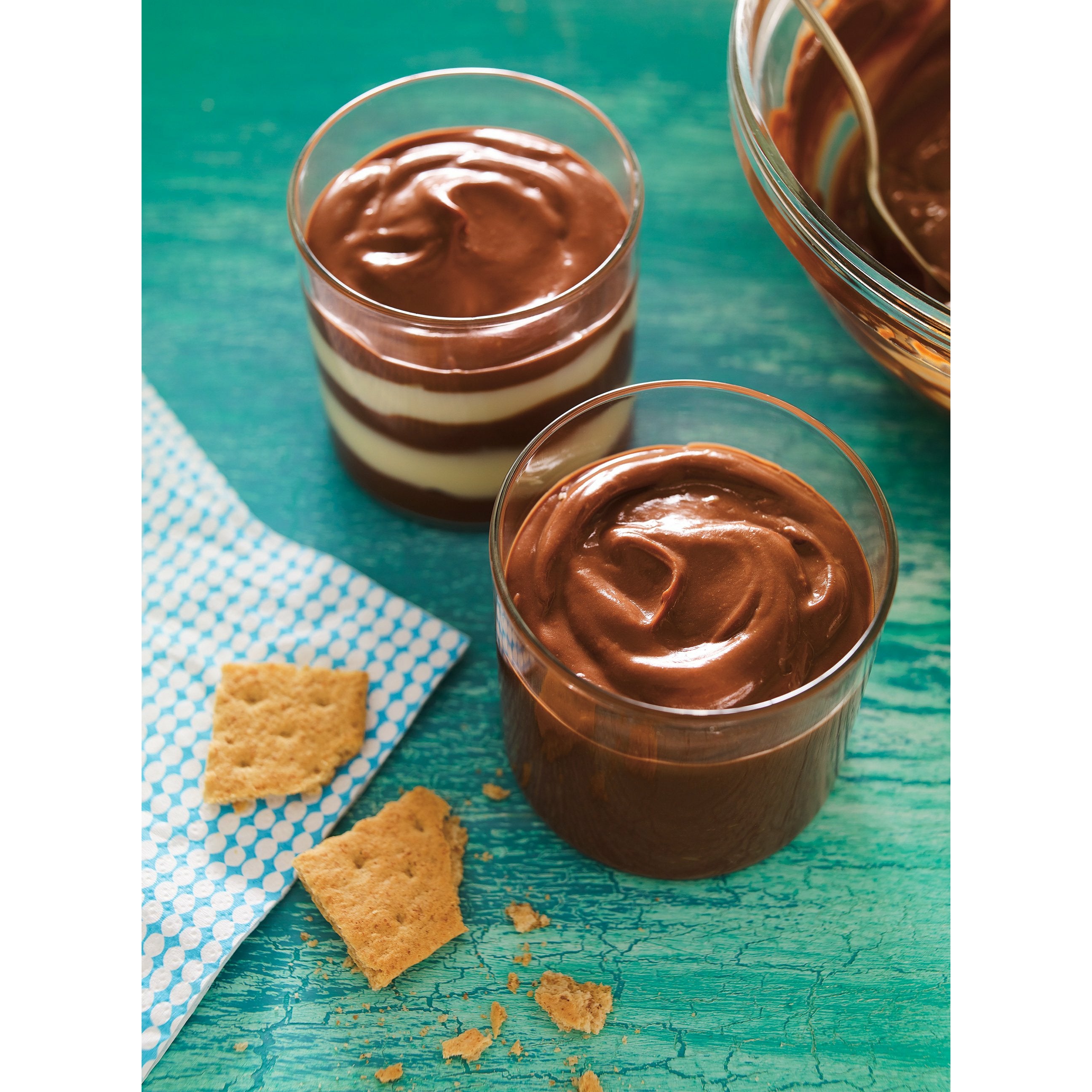 2 verres transparents remplis d’un pudding couleur crème, à droite il y a un bol transparent de crème au chocolat. Le tout sur une table bleu-vert