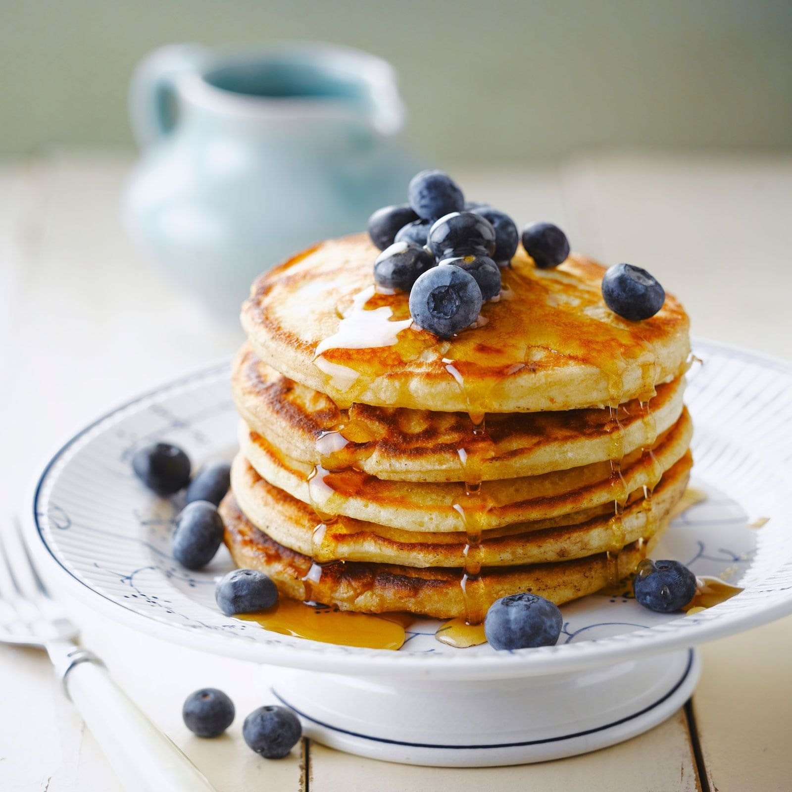 Une assiette blanche avec des pancakes, du sirop brun et des myrtilles et à l’arrière une petite carafe bleu. Le tout sur une table en bois blanche