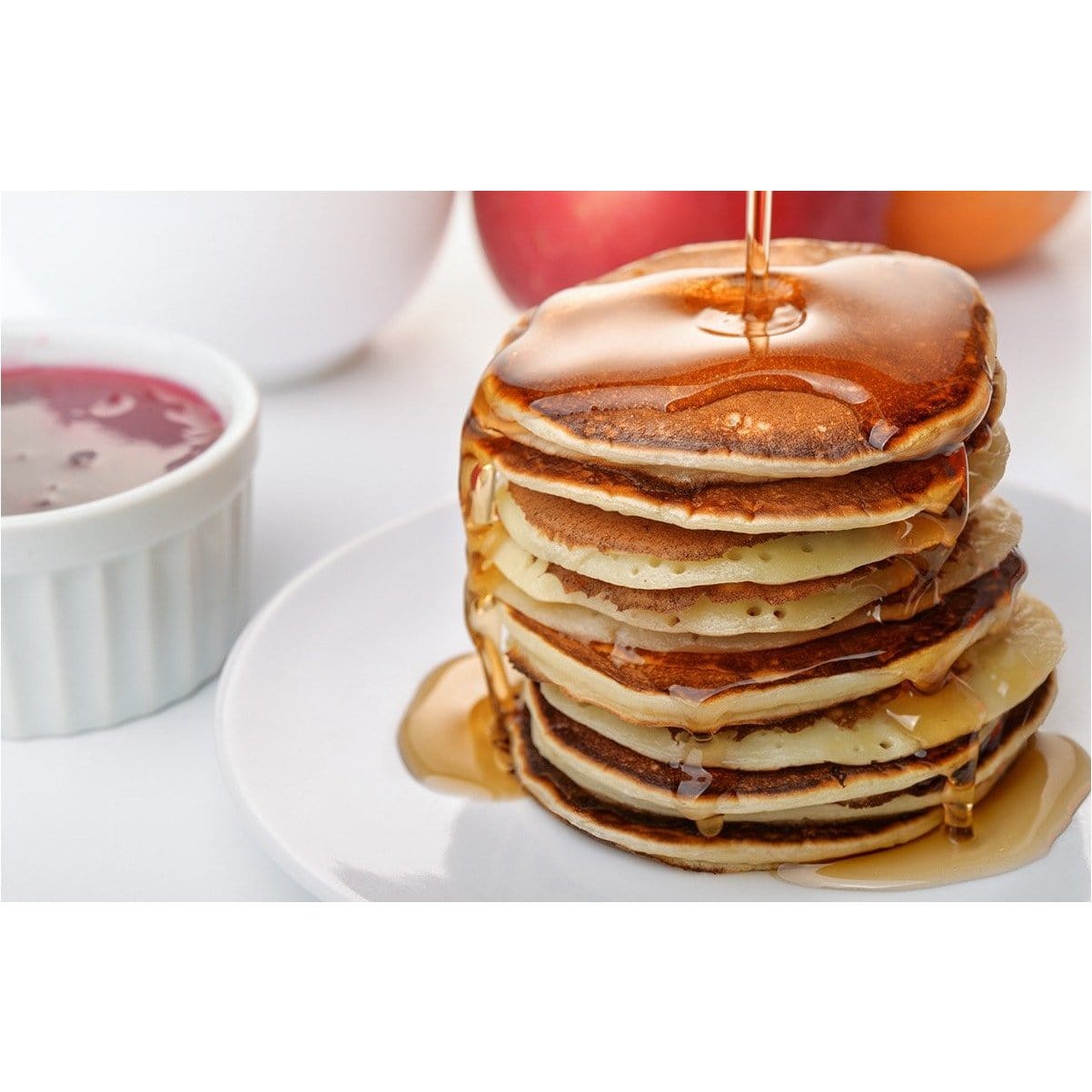 Une assiette blanche avec des pancakes, du sirop brun et un petit bol blanc avec une confiture rouge. Le tout sur une table blanche