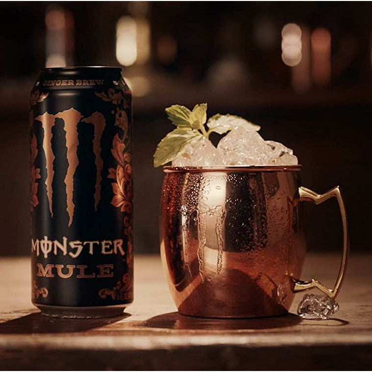 Une grande canette noire avec au centre le logo rose saumon de Monster, un grand M et sa droite il y a une petite tasse rose saumon. Le tout sur une table en bois
