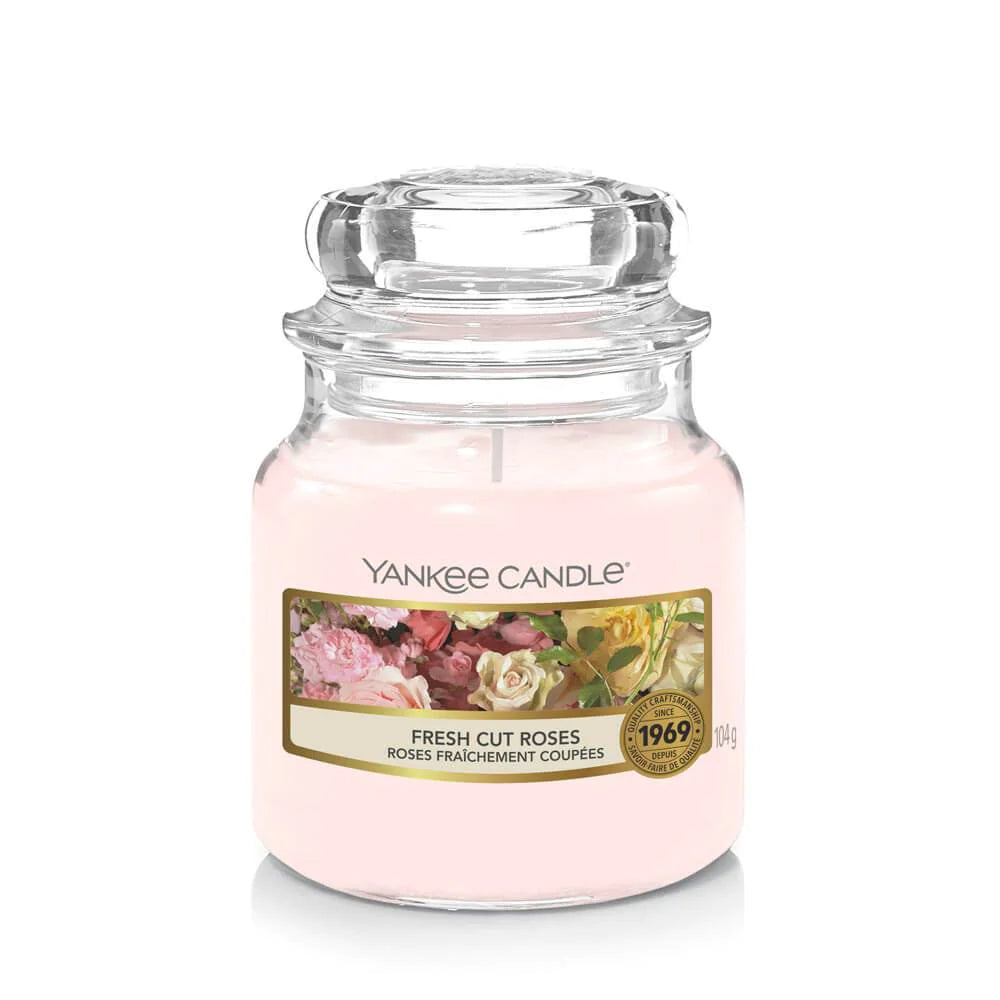 Un emballage sur fond blanc avec une bougie rose pâle dans un bocal en verre, un capuchon en verre et une étiquette avec plusieurs rosses blanches, roses, rouges