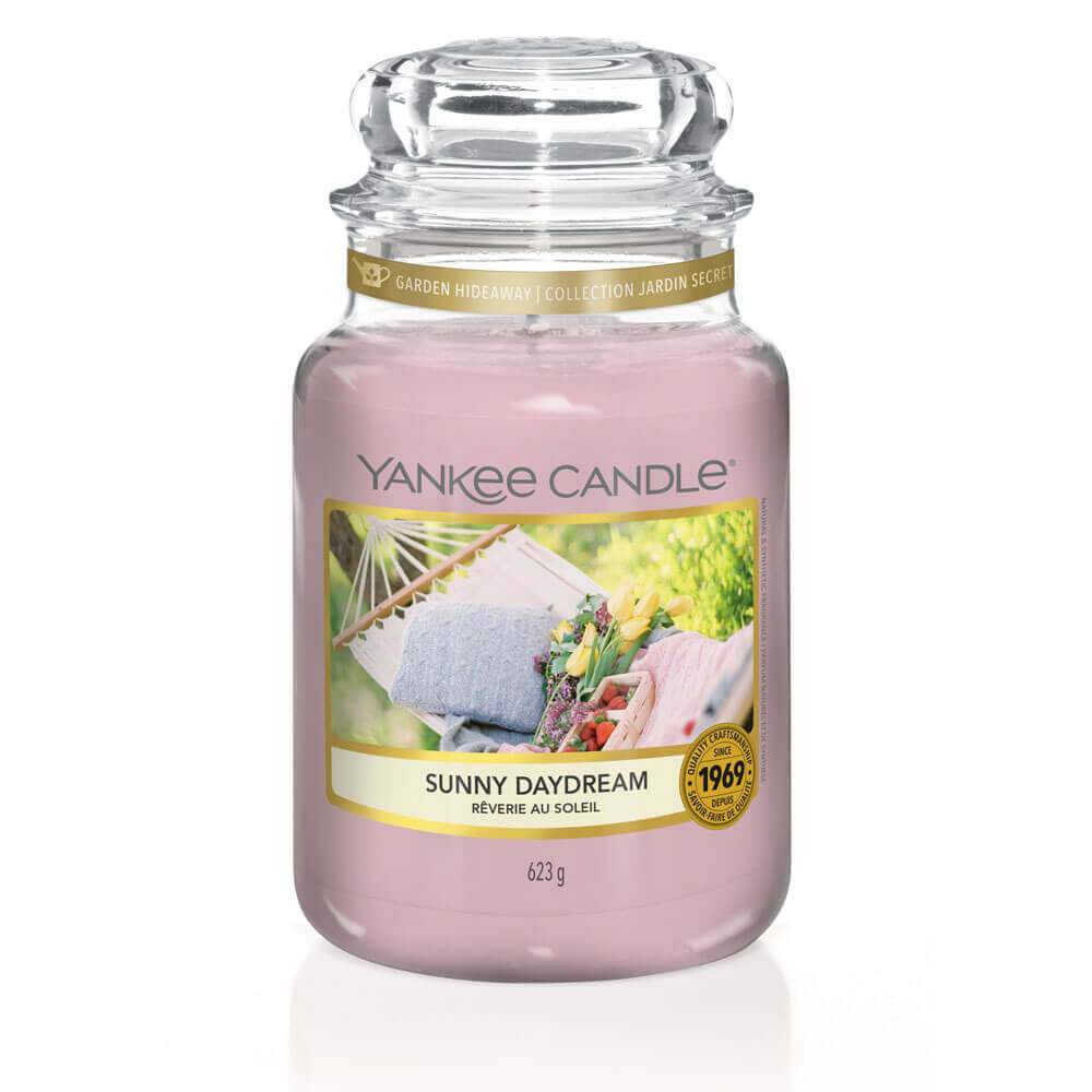 Un emballage sur fond blanc avec une bougie rose dans un bocal en verre, un capuchon en verre et une étiquette avec des tissus et des fleurs sur un hamac blanc dans un jardin